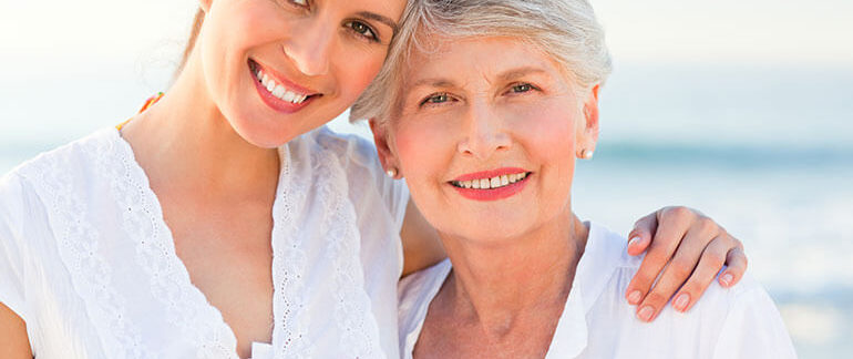 A woman and her mother smile for a picture on the beach.