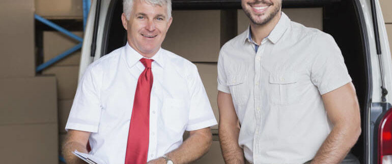 Two business partners stand in front of a car and warehouse full of boxes