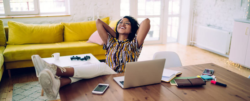 A woman is sitting at her table in front of her laptop, relaxing with her hands behind her head.
