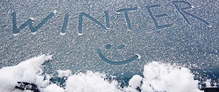 A car's window has the word "Winter" spelled out in the snow that has accumulated on top of it. There is also a smiley face drawn below the word.