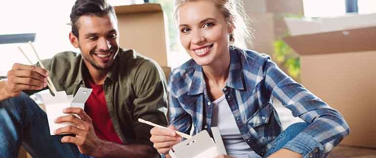 Man and woman sitting on the floor eating out of take out containers.