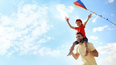 Young boy sitting on father's shoulders while flying a kite.