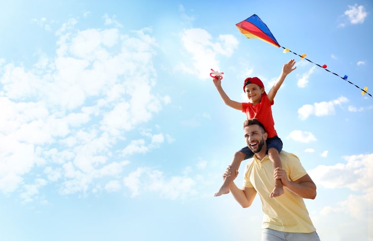 Young boy sitting on father's shoulders while flying a kite.