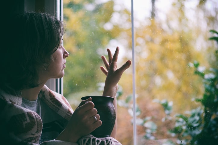 A woman is wrapped in a blanket beside a window, holding a mug in one hand and pressing the other to the glass.