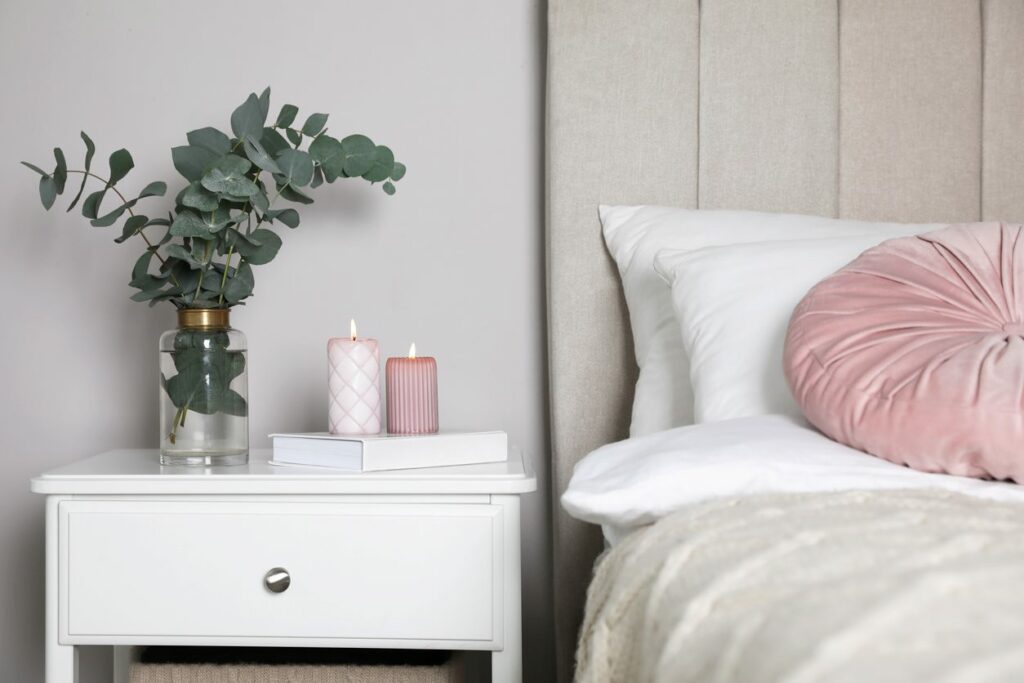A white nightstand is decorated with foliage, candles, and a book next to a bed.