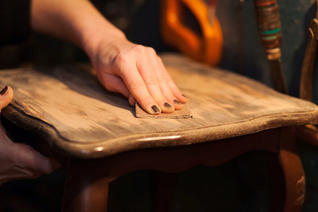 Hands use sandpaper to smooth the surface of a wooden chair.