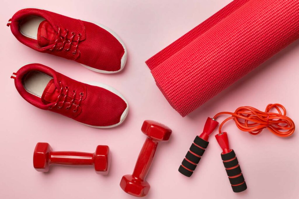 Red tennis shoes, red yoga mat, red dumbbells, and red jump rope on a pink background