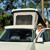 A woman waves to the camera, about to enter her camper van