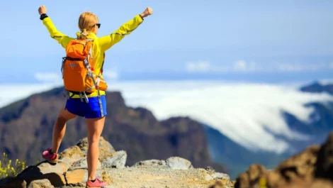 A woman in blue shorts and a yellow hiking jacket celebrates at the top of a long trail