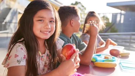 Three children sitting at a lunch table talking to each other while eating