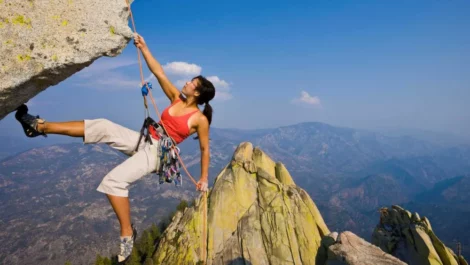 A female rock climber wearing a red tank top hangs from a high cliff