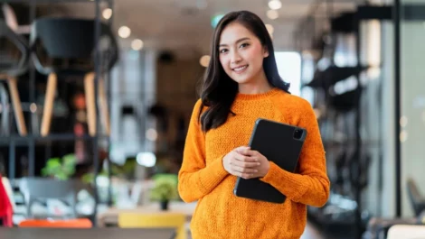A smiling businesswoman stands in a workspace holding a tablet