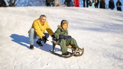 Father smiles while pushing his sun down on a snowy hill on a sled.