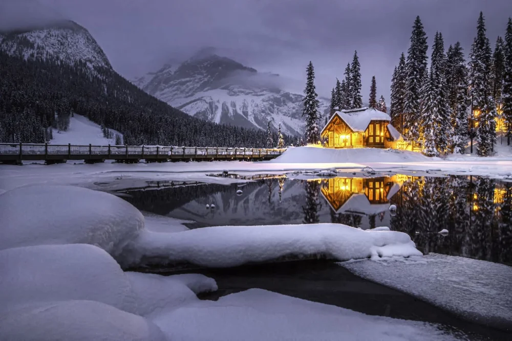 A cozy wooden cabin glows with yellow lights in a snowy mountainside forest near a lake
