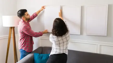 A young couple hangs a plain white canvas on the wall next to two other plain canvases.