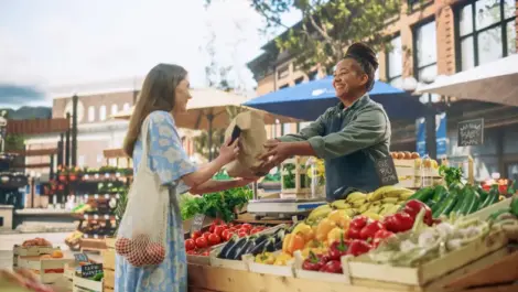 A smiling vendor hands a lady in a blue dress a paper bag of produce at a farmers market